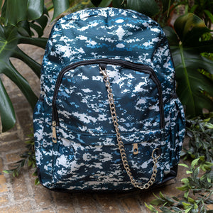 The Pixel Camouflage Backpack sat outside on a brick floor surrounded by giant monstera leaves and other green foliage. The navy, blue and white vegan friendly backpack is sat facing forward to highlight the two front zip pockets with a silver draping detachable chain, the two side elasticated pockets, the top handle and the main double zip compartment.