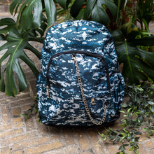 Load image into Gallery viewer, The Pixel Camouflage Backpack sat outside on a brick floor surrounded by giant monstera leaves and other green foliage. The navy, blue and white vegan friendly backpack is sat facing forward to highlight the two front zip pockets with a silver draping detachable chain, the two side elasticated pockets, the top handle and the main double zip compartment.