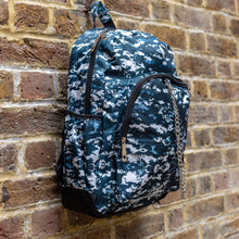 Load image into Gallery viewer, The Pixel Camouflage Backpack hanging outside on a brick wall. The navy, blue and white vegan friendly backpack is facing forward to highlight the two front zip pockets with a silver draping detachable chain, the two side elasticated pockets, the top handle and the main double zip compartment.
