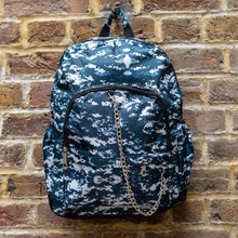 Load image into Gallery viewer, The Pixel Camouflage Backpack hanging outside on a brick wall. The navy, blue and white vegan friendly backpack is facing forward to highlight the two front zip pockets with a silver draping detachable chain, the two side elasticated pockets, the top handle and the main double zip compartment.