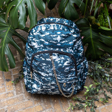 Load image into Gallery viewer, The Pixel Camouflage Backpack sat outside on a brick floor surrounded by giant monstera leaves and other green foliage. The navy, blue and white vegan friendly backpack is sat facing forward to highlight the two front zip pockets with a silver draping detachable chain, the two side elasticated pockets, the top handle and the main double zip compartment.