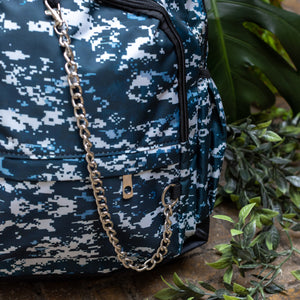 Close up of The Pixel Camouflage Backpack sat outside on a brick floor surrounded by giant monstera leaves and other green foliage. The navy, blue and white vegan friendly backpack is sat facing forward to highlight the two front zip pockets with a silver draping detachable chain, the two side elasticated pockets, the top handle and the main double zip compartment.