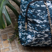 Load image into Gallery viewer, Close up of The Pixel Camouflage Backpack sat outside on a brick floor surrounded by giant monstera leaves and other green foliage. The navy, blue and white vegan friendly backpack is sat facing forward to highlight the two front zip pockets with a silver draping detachable chain, the two side elasticated pockets, the top handle and the main double zip compartment.
