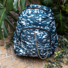 Load image into Gallery viewer, The Pixel Camouflage Backpack sat outside on a brick floor surrounded by giant monstera leaves and other green foliage. The navy, blue and white vegan friendly backpack is sat facing forward to highlight the two front zip pockets with a silver draping detachable chain, the two side elasticated pockets, the top handle and the main double zip compartment.
