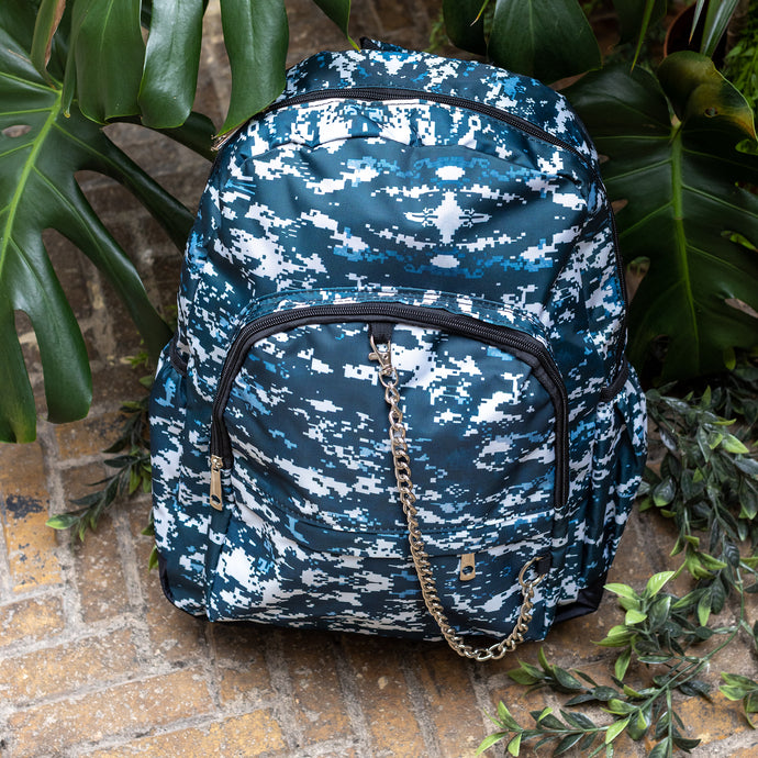 The Pixel Camouflage Backpack sat outside on a brick floor surrounded by giant monstera leaves and other green foliage. The navy, blue and white vegan friendly backpack is sat facing forward to highlight the two front zip pockets with a silver draping detachable chain, the two side elasticated pockets, the top handle and the main double zip compartment.