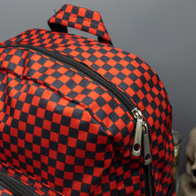 Load image into Gallery viewer, Close up of the Red Checkerboard Backpack sat on a grey background with a palmistry guide book and two skull stack on the right and a phrenology guide candle and three skull stack on the left. The vegan friendly bag is facing forward to highlight the red and black check print, two front zip pockets, two elasticated side pockets, main top double zip pocket and silver draping decorative chain.