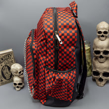 Load image into Gallery viewer, The Red Checkerboard Backpack sat on a grey background with a palmistry guide book and two skull stack on the right and a phrenology guide candle and three skull stack on the left. The vegan friendly bag is facing left to highlight the red and black check print, two front zip pockets, two elasticated side pockets, main top double zip pocket and silver draping decorative chain.