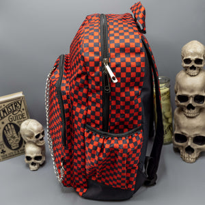 The Red Checkerboard Backpack sat on a grey background with a palmistry guide book and two skull stack on the right and a phrenology guide candle and three skull stack on the left. The vegan friendly bag is facing left to highlight the red and black check print, two front zip pockets, two elasticated side pockets, main top double zip pocket and silver draping decorative chain.