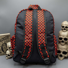 Load image into Gallery viewer, The Red Checkerboard Backpack sat on a grey background with a palmistry guide book and two skull stack on the right and a phrenology guide candle and three skull stack on the left. The vegan friendly bag is facing away from the camera to highlight the plain black back, the two side elasticated pockets, the top handle and the two adjustable padded shoulder straps.