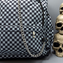 Load image into Gallery viewer, Close up of the Grey Checkerboard Backpack sat on a grey background with a palmistry guide book and two skull stack on the right and a phrenology guide candle and three skull stack on the left. The vegan friendly bag is facing forward to highlight the red and black check print, two front zip pockets, two elasticated side pockets, main top double zip pocket and silver draping decorative chain.

