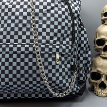 Load image into Gallery viewer, Close up of the Grey Checkerboard Backpack sat on a grey background with a palmistry guide book and two skull stack on the right and a phrenology guide candle and three skull stack on the left. The vegan friendly bag is facing forward to highlight the red and black check print, two front zip pockets, two elasticated side pockets, main top double zip pocket and silver draping decorative chain.