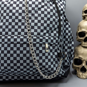 Close up of the Grey Checkerboard Backpack sat on a grey background with a palmistry guide book and two skull stack on the right and a phrenology guide candle and three skull stack on the left. The vegan friendly bag is facing forward to highlight the red and black check print, two front zip pockets, two elasticated side pockets, main top double zip pocket and silver draping decorative chain.
