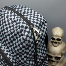 Load image into Gallery viewer, Close up of the Grey Checkerboard Backpack sat on a grey background with a palmistry guide book and two skull stack on the right and a phrenology guide candle and three skull stack on the left. The vegan friendly bag is facing forward to highlight the grey and black check print, two front zip pockets, two elasticated side pockets, main top double zip pocket and silver draping decorative chain.
