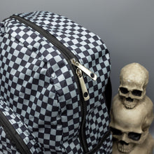 Load image into Gallery viewer, Close up of the Grey Checkerboard Backpack sat on a grey background with a palmistry guide book and two skull stack on the right and a phrenology guide candle and three skull stack on the left. The vegan friendly bag is facing forward to highlight the grey and black check print, two front zip pockets, two elasticated side pockets, main top double zip pocket and silver draping decorative chain.