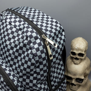 Close up of the Grey Checkerboard Backpack sat on a grey background with a palmistry guide book and two skull stack on the right and a phrenology guide candle and three skull stack on the left. The vegan friendly bag is facing forward to highlight the grey and black check print, two front zip pockets, two elasticated side pockets, main top double zip pocket and silver draping decorative chain.