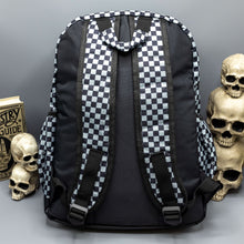 Load image into Gallery viewer,  The Grey Checkerboard Backpack sat on a grey background with a palmistry guide book and two skull stack on the right and a phrenology guide candle and three skull stack on the left. The vegan friendly bag is facing away from the camera to highlight the plain black back, the two side elasticated pockets, the top handle and the two adjustable padded shoulder straps.