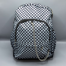 Load image into Gallery viewer, The Grey Checkerboard Backpack sat on a grey background with a palmistry guide book and two skull stack on the right and a phrenology guide candle and three skull stack on the left. The vegan friendly bag is facing forward to highlight the red and black check print, two front zip pockets, two elasticated side pockets, main top double zip pocket and silver draping decorative chain.

