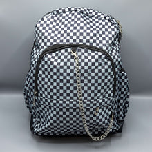 Load image into Gallery viewer, The Grey Checkerboard Backpack sat on a grey background with a palmistry guide book and two skull stack on the right and a phrenology guide candle and three skull stack on the left. The vegan friendly bag is facing forward to highlight the red and black check print, two front zip pockets, two elasticated side pockets, main top double zip pocket and silver draping decorative chain.