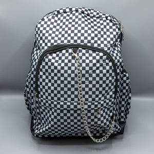 The Grey Checkerboard Backpack sat on a grey background with a palmistry guide book and two skull stack on the right and a phrenology guide candle and three skull stack on the left. The vegan friendly bag is facing forward to highlight the red and black check print, two front zip pockets, two elasticated side pockets, main top double zip pocket and silver draping decorative chain.