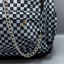 Load image into Gallery viewer, Close up of the Grey Checkerboard Backpack sat on a grey background. The vegan friendly bag is facing forward to highlight the grey and black check print, two front zip pockets, two elasticated side pockets, main top double zip pocket and silver draping decorative chain.
