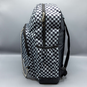  The Grey Checkerboard Backpack sat on a grey background. The vegan friendly bag is facing left to highlight the grey and black check print, two front zip pockets, two elasticated side pockets, main top double zip pocket and silver draping decorative chain.