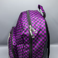 Load image into Gallery viewer, Close up of the Purple Checkerboard Backpack sat on a grey background. The vegan friendly bag is facing forward to highlight the purple and black check print, two front zip pockets, two elasticated side pockets, main top double zip pocket and silver draping decorative chain.