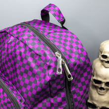Load image into Gallery viewer, Close up of the Purple Checkerboard Backpack sat on a grey background with a palmistry guide book and two skull stack on the right and a phrenology guide candle and three skull stack on the left. The vegan friendly bag is facing forward to highlight the purple and black check print, two front zip pockets, two elasticated side pockets, main top double zip pocket and silver draping decorative chain.