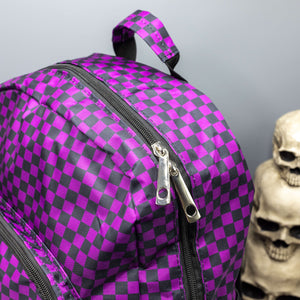 Close up of the Purple Checkerboard Backpack sat on a grey background with a palmistry guide book and two skull stack on the right and a phrenology guide candle and three skull stack on the left. The vegan friendly bag is facing forward to highlight the purple and black check print, two front zip pockets, two elasticated side pockets, main top double zip pocket and silver draping decorative chain.