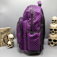 Load image into Gallery viewer, The Purple Checkerboard Backpack sat on a grey background with a palmistry guide book and two skull stack on the right and a phrenology guide candle and three skull stack on the left. The vegan friendly bag is facing left to highlight the purple and black check print, two front zip pockets, two elasticated side pockets, main top double zip pocket and silver draping decorative chain.