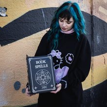 Load image into Gallery viewer, A blue haired model stood in front of a graffiti wall wearing an oversized black jumper whilst holding the GothX book of spells vegan messenger bag down by their side. Vegan black leather with white witch pagan magic symbols printed on the front and spine of the 3d book bag.