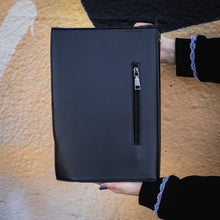 Load image into Gallery viewer, The GothX Book of Spells Vegan Messenger Bag. Vegan black leather with white witch pagan magic symbols printed on the front and spine of the 3d book bag. Bag held up in front of a graffiti wall to show off the back zip up pocket.