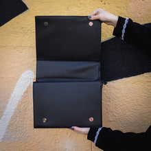 Load image into Gallery viewer, The GothX Book of Spells Vegan Messenger Bag. Vegan black leather with white witch pagan magic symbols printed on the front and spine of the 3d book bag. Bag held up in front of a graffiti wall to show off the inside button close and zip up features.