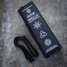 Load image into Gallery viewer, The GothX Book of Spells Vegan Messenger Bag. Vegan black leather with white witch pagan magic symbols printed on the front and spine of the 3d book bag. The book of spells bag is laying on its side to show off the magical symbol printed spine and the detachable adjustable strap laying next to it on a brick bench.