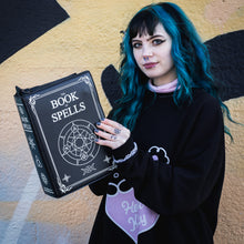 Load image into Gallery viewer, A blue haired model stood in front of a graffiti wall wearing an oversized black jumper whilst holding the GothX book of spells vegan messenger bag up as if they're reading it. Vegan black leather with white witch pagan magic symbols printed on the front and spine of the 3d book bag.