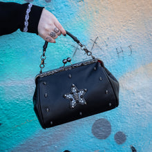 Load image into Gallery viewer, The GothX skull struck vegan vintage clasp handbag being held up in front of a graffiti wall. The vegan leather vintage goth inspired bag is facing forward to highlight the vintage ball clasp close, floral metal top detailing, detachable handle strap, skull studs and skull star with chains, studs and crystals.