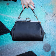 Load image into Gallery viewer, The GothX skull struck vegan vintage clasp handbag being held up in front of a graffiti wall. The bag is facing away from the camera to highlight the vintage ball clasp close and detachable handle strap.