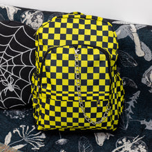 Load image into Gallery viewer, The CHOK yellow checker vegan backpack is sat on a spooky halloween blanket next to a spiderweb cushion facing forward. The backpack is yellow and black all over checkerboard with two front black zip pockets, two side pockets and a silver decorative chain going across the front.