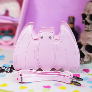 The GothX Pastel Pink Bat Vegan Shoulder Bag on a pastel purple background with multicoloured confetti, pastel pink coffin shelving and black skull bottles surrounding it. The bag is facing forward to highlight the embroidered detailing, crystal eyes and detachable adjustable strap.