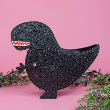 Load image into Gallery viewer, The kawaii black glitter dino vegan bag on a pink studio background with green foliage surrounding it. The bag is facing forward to highlight the dinosaur face, moveable arm, detachable strap and black glitter front.