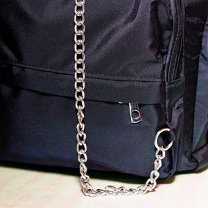 The black nylon vegan backpack with chain on an iridescent background. Close up of the detachable silver chain and front zip pocket detailing.