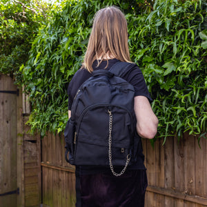 Jack is stood outside in a garden area wearing the black nylon vegan backpack with chain on his back whilst facing away from the camera. The photo is cropped from the thighs up. The backpack is all black with two front zip pockets, two side pockets and a silver decorative chain across the front to the side.