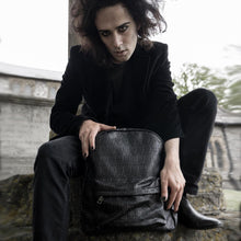 Load image into Gallery viewer, Sweeney Deville sat near a graveyard in an all black gothic outfit posing with the gothx embossed skull vegan backpack whilst sat down and leaning forward.