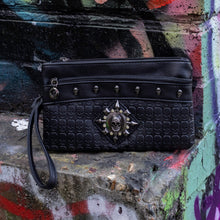 Load image into Gallery viewer, The gothx black skull vegan clutch bag on an alternative colourful graffiti brick wall outside. The vegan clutch bag is facing forward to highlight the skull embossed vegan black leather, the crystal stud skull centrepiece, zip pocket and skull studs.