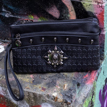 Load image into Gallery viewer, The gothx black skull vegan clutch bag on an alternative colourful graffiti brick wall outside. The vegan clutch bag is facing forward to highlight the skull embossed vegan black leather, the crystal stud skull centrepiece, zip pocket and skull studs.