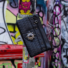 Load image into Gallery viewer, The gothx black skull vegan clutch bag on an alternative colourful graffiti brick wall outside. The vegan clutch bag is facing forward to highlight the skull embossed vegan black leather, the crystal stud skull centrepiece, zip pocket and skull studs.