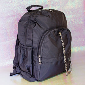The black nylon vegan backpack with chain on an iridescent background. The bag is facing forward angled right to highlight the two front zip pockets, two side pockets, double zip main compartment, top handle and detachable silver chain.