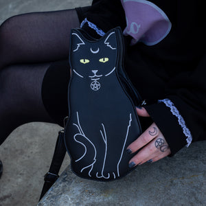 The gothx pagan black cat vegan shoulder bag resting next to a model wearing an all black hello kitty outfit. The bag is facing forward to highlight the embroidery details including a pentagram collar, yellow eyes and paws.