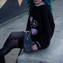 Load image into Gallery viewer, Model with blue hair wearing a hello kitty outfit holding the gothx pagan black cat vegan shoulder bag next to them. The bag is facing forward to highlight the embroidery details including a pentagram collar, yellow eyes and paws.
