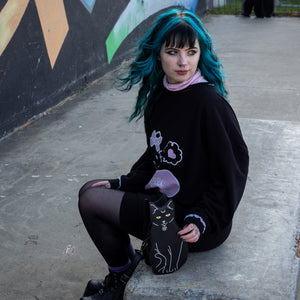 Model with blue hair wearing a hello kitty outfit holding the gothx pagan black cat vegan shoulder bag next to them. The bag is facing forward to highlight the embroidery details including a pentagram collar, yellow eyes and paws.