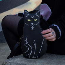 Load image into Gallery viewer, The gothx pagan black cat vegan shoulder bag resting next to a model wearing an all black hello kitty outfit. The bag is facing forward to highlight the embroidery details including a pentagram collar, yellow eyes and paws.
